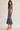 A woman modeling a blue floral midi dress with spaghetti straps, a cowl neckline, and a flared ruffled hem, paired with clear high-heeled sandals against a plain light beige studio background.