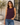 A woman wearing a sleeveless crewneck tank top in deep plum purple and blue jeans, standing outdoors in front of a modern building with a potted tree.