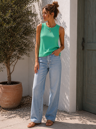 Woman wearing a green tank top and blue jeans standing against a white wall with a plant in the background.