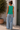 Woman wearing a green sleeveless top and blue jeans standing outdoors.
