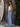 Woman in a white top and blue pants standing next to a wooden wall with a surfboard.