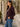 A woman wearing a dark plum purple blazer over a white top and blue jeans, standing outdoors against a textured stucco wall with green foliage.