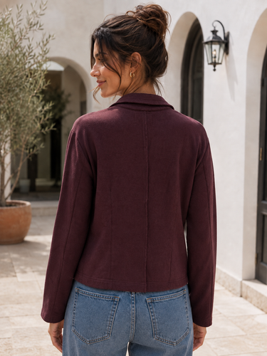 A woman wearing a long-sleeve cowl-neck jacket in deep burgundy, shown from the back. She is wearing blue jeans and standing in an outdoor courtyard with a white stucco wall, arches, and a potted olive tree in the background.