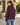 A woman wearing a dark burgundy long-sleeve cropped jacket and blue jeans, photographed from behind in an outdoor courtyard setting.