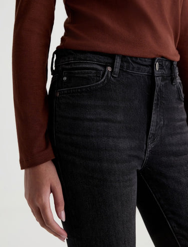 A woman wearing slim straight jeans in black with black boots and a maroon long sleeve tee on a white background.