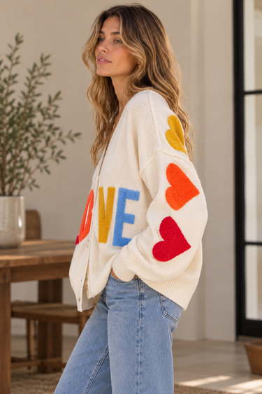 A woman wearing a beige v-neck cardigan with “LOVE” written in multi-colored letters at the middle of the sweater and hearts down the sleeve