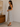 Woman wearing a brown top and patterned skirt standing in a room with a wooden dresser and framed picture.
