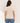 A woman viewed from behind wearing a fitted short-sleeve crew-neck t-shirt in a light cream color, paired with blue jeans, against a plain off-white wall.