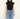 A woman wearing a black sleeveless mock-neck top and light blue jeans, standing against a plain off-white background.