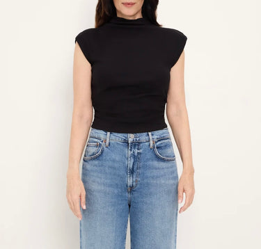A woman wearing a black sleeveless mock-neck top and light blue jeans, standing against a plain off-white background.