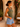 A woman wearing a sage green ruched tank top and light blue denim shorts, standing in a sunlit outdoor patio setting with a wicker chair and potted plants.