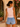 Woman in a light pink tank top and denim shorts standing outdoors with flowers and a chair in the background.