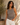 Woman wearing a gray tank top and white pants standing against a textured wall with a potted plant.