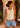 A woman wearing a cream ribbed tank top and light blue denim shorts, standing on a sunlit patio with a stucco wall, potted plants, and a wicker chair in the background.