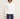 A woman wearing a white long-sleeve knit top with a wide boat neckline and textured vertical ribbing, paired with dark blue jeans, against a plain light beige background.