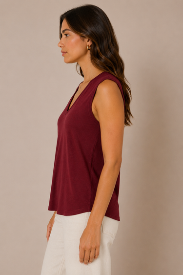 A woman shown in side profile wearing a sleeveless V-neck top in deep burgundy and white pants, standing against a plain warm beige studio background.