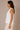 A woman shown in side profile wearing a white sleeveless V-neck top and light beige pants against a plain beige studio background.