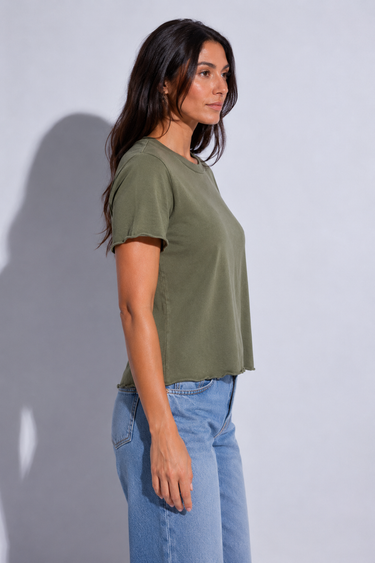 A woman wearing a short-sleeve crewneck t-shirt in olive green, paired with light wash blue jeans, standing in profile against a light gray studio background.