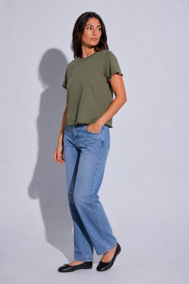 A woman modeling a short-sleeve olive green t-shirt and light wash blue jeans, standing against a light gray studio background with a soft shadow.