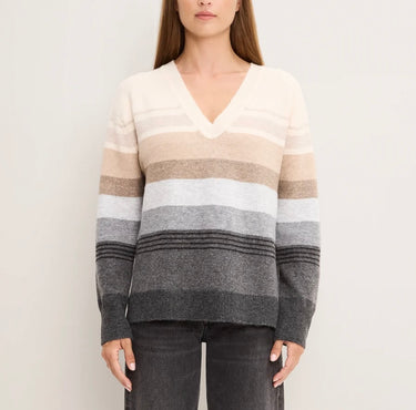 A woman wearing a long-sleeve V-neck knit sweater with horizontal stripes in cream, beige, taupe, light gray, medium gray, and charcoal, paired with dark gray jeans against a plain off-white background.