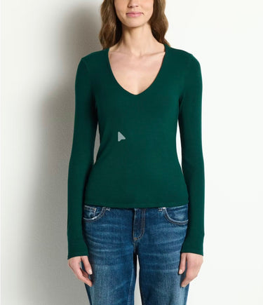 A woman wearing a dark green long-sleeve V-neck knit top with a small white geometric logo on the chest, paired with blue jeans, standing against a plain off-white wall.