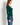 A woman wearing a dark green long-sleeve V-neck top, paired with blue jeans, standing against a plain off-white wall.