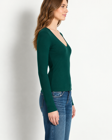 A woman wearing a dark green long-sleeve V-neck top, paired with blue jeans, standing against a plain off-white wall.