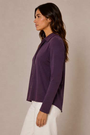 A woman in side profile wearing a long-sleeve button-up shirt in a deep purple color, paired with white pants, against a plain beige studio background.