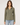 A woman modeling a long-sleeve button-up shirt in olive green, paired with light-wash blue jeans, against a plain light gray studio background.