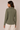 A woman shown from the back wearing an olive green long-sleeve button-up shirt and cream pants, standing against a plain beige studio background.