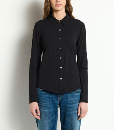 A woman wearing a black long-sleeve button-up shirt and blue jeans, standing against a plain off-white wall.