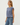 A woman wearing a dusty blue sleeveless top and blue jeans, shown from a side profile against a plain light gray studio background.