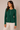 A woman wearing a dark green long-sleeve ribbed knit top with a buttoned neckline and cream pants, standing against a plain beige studio background.