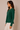 A woman wearing a dark green long-sleeve knit top with decorative white buttons down the front, paired with white pants, standing against a plain beige studio background.