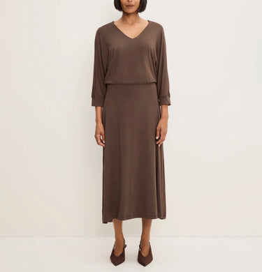 A woman modeling a solid brown midi dress with a V-neckline, three-quarter sleeves, and a draped overlay, paired with brown pointed-toe heels against a plain off-white studio background.