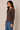 A woman wearing a dark brown long-sleeve button-front cardigan and blue jeans, standing against a plain beige studio background.