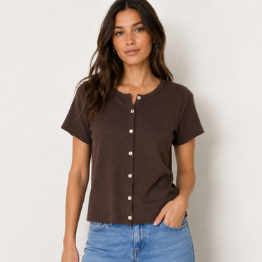 A woman wearing a dark brown short-sleeve henley top with white buttons, paired with blue jeans, standing against a plain off-white wall.