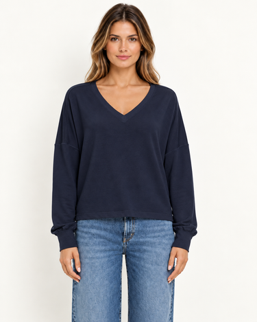 A woman facing foreward wearing a navy v-neck long sleeve and blue jeans on a white background.