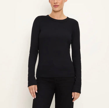 A woman wearing a black long-sleeve crewneck top and black pants, standing against a plain off-white background.