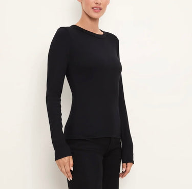 A woman wearing a fitted black long-sleeve crewneck top and black pants, standing against a plain off-white studio background.