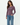 A woman modeling a long-sleeve crewneck knit top in a dusty mauve-purple color, paired with blue jeans, against a clean white studio background.