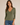 A woman modeling a fitted olive green long-sleeve V-neck top with three-quarter sleeves, paired with blue jeans, against a plain beige studio background.