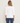 A woman viewed from the back wearing a white three-quarter sleeve top and blue jeans, standing against a plain off-white studio background.
