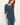 A woman wearing a dark teal ribbed 3/4-sleeve scoop-neck top and blue jeans, standing against a plain off-white background.