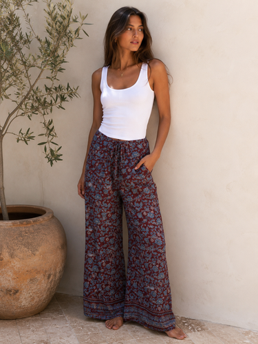 Woman wearing a white tank top and patterned pants standing against a neutral wall with a plant in the background.