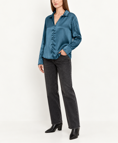 A woman modeling a teal blue satin button-up shirt with ruffle details down the front, paired with dark gray jeans and black ankle boots, against a plain white studio background.