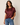 A woman wearing a short-sleeve crewneck satin top in a deep burgundy color, paired with blue jeans, standing against a plain light beige background.