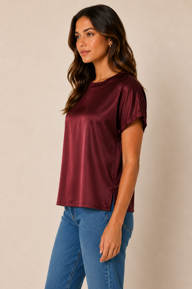 A woman wearing a short-sleeve satin top in deep burgundy with blue jeans, standing against a plain beige studio background.
