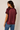 A woman shown from the back wearing a short-sleeve satin-like top in deep burgundy with contrasting darker burgundy raglan sleeves, paired with blue jeans, against a plain light beige studio background.