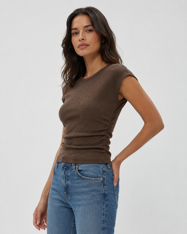 A woman modeling a brown ribbed short-sleeve top with side ruching, paired with blue jeans, against a plain light gray studio background.